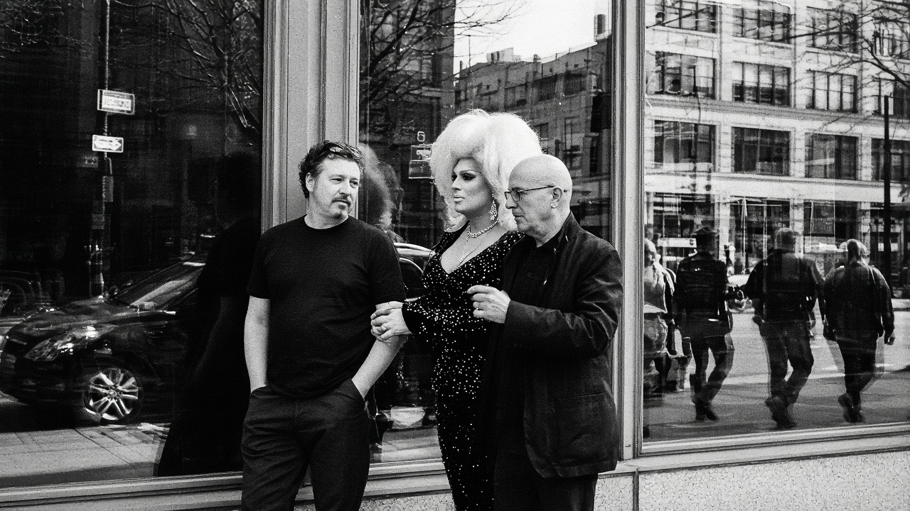Drag queen with a voluminous bouffant wig stands between two men outside a downtown storefront window