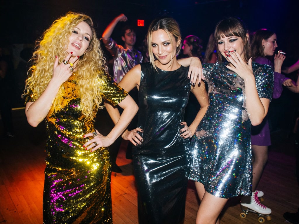 Three women in sparkly party dresses pose together on a retro disco club dance floor