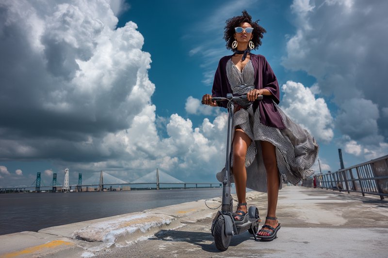 Bold graphic linework with confident contour definition combined with luminous plein-air color washes and dramatic atmospheric skies, creating a vivid editorial illustration fusion. The result should manifest as crisp figure delineation against painterly cumulus cloudscapes, with saturated naturalistic tones, strong directional light, and a cinematic low-angle perspective that heroizes the subject.

A curly-haired woman in dark sunglasses and hoop earrings, wearing a flowing grey sundress and deep purple open cardigan with flip-flops, standing confidently astride an electric scooter, hands gripping the handlebars, chin lifted, gazing into the distance, positioned left of center and towering into the frame.

Scene set on a wide concrete waterfront promenade during a bright breezy summer afternoon, with a metal railing, river glinting below, a distant steel lift-bridge on the right horizon, and a vast sky filled with billowing cumulus clouds, shot from a dramatic low ground-level camera angle looking upward.