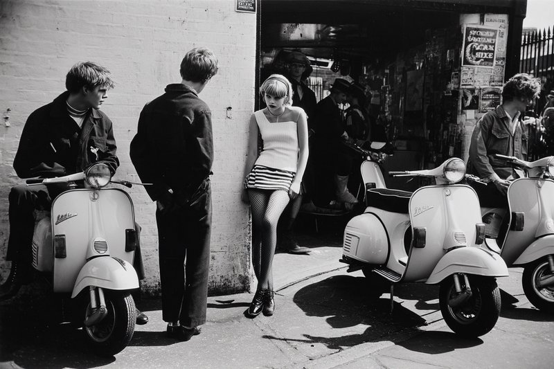 Black-and-white street scene with a stylish woman leaning against a wall beside vintage scooters.