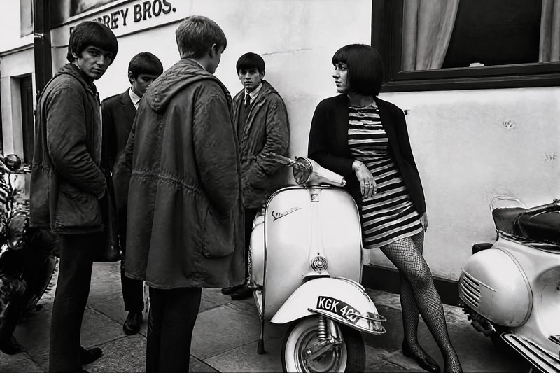 Black-and-white street scene with a stylish woman leaning on a vintage Vespa while a group of young men chat nearby.