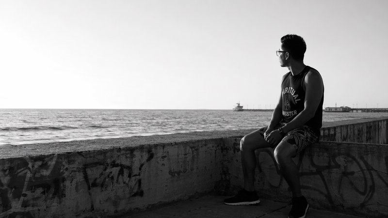 A man with glasses sits on a graffiti-covered concrete seawall, gazing out at a calm ocean in monochrome.