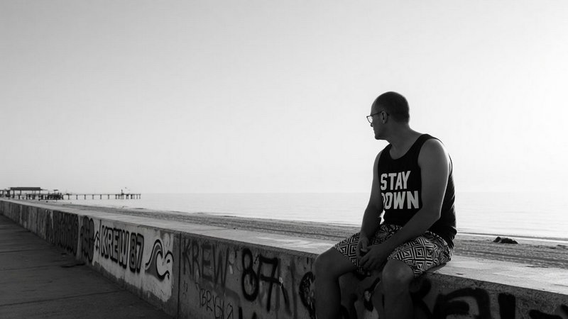 [Monochrome shot of a man sitting on a graffiti-covered seawall, looking out over a calm beach and ocean]