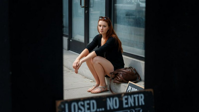 Pensive young red-haired woman sits on storefront steps, viewed through a doorway frame with a closed sign in the foreground