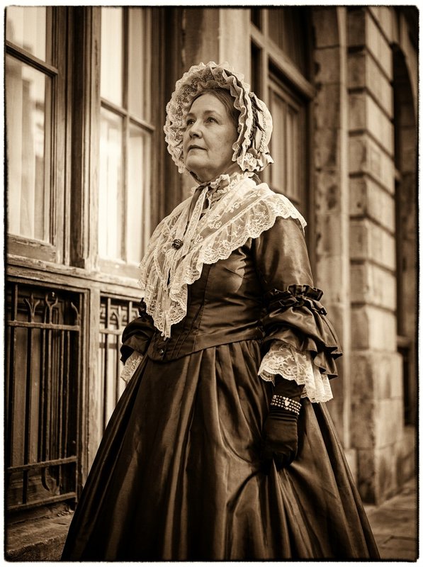 Sepia portrait of a woman in elaborate Victorian period dress and lace bonnet standing outside a historic stone building