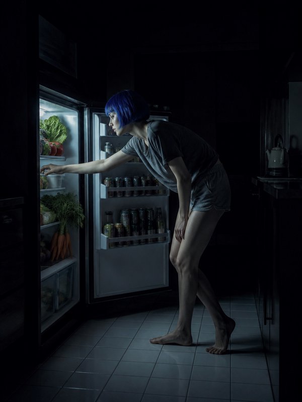 Woman with short blue hair reaches into an illuminated open refrigerator in a dark midnight kitchen