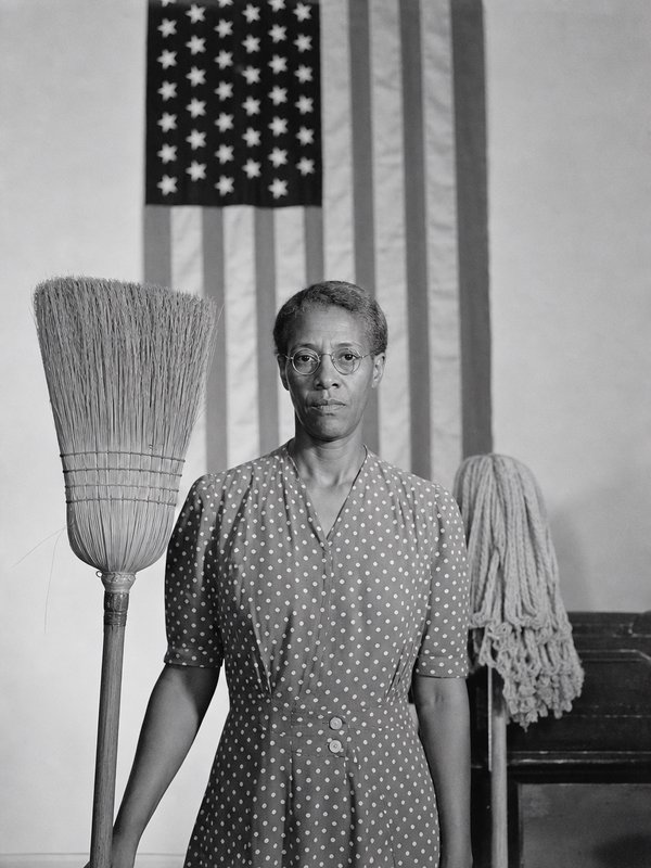 Dignified Black domestic worker holds straw broom, standing before a US flag in this vintage black and white portrait.