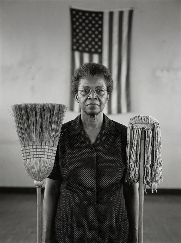 Black and white portrait of a dignified Black female custodian standing between a broom and mop, US flag on wall behind her.