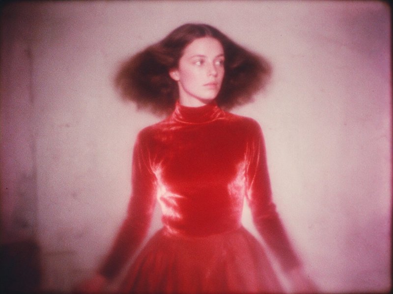 Soft focus vintage film portrait of a woman in a red velvet dress, dark hair fanned out against a hazy pale pink background.