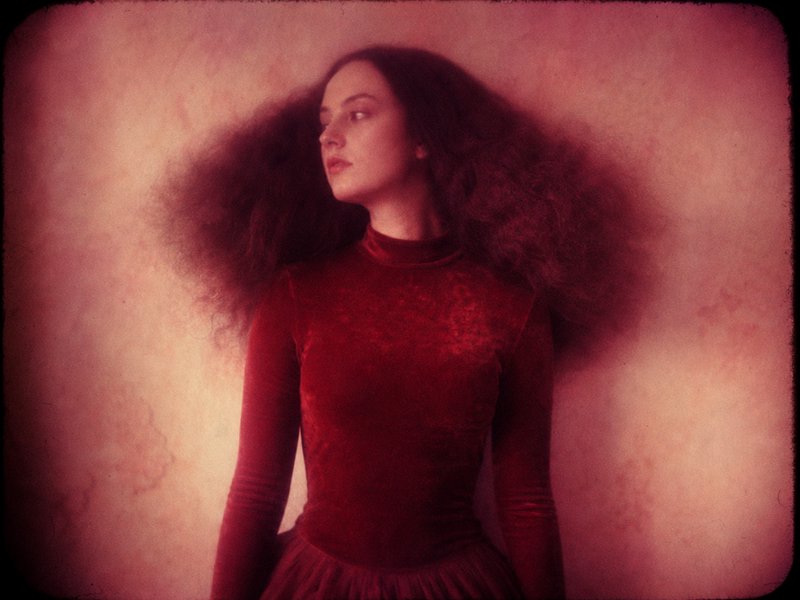 [Soft focus vintage film portrait of a woman with voluminous curly hair in a red velvet dress, gazing left against a warm ...