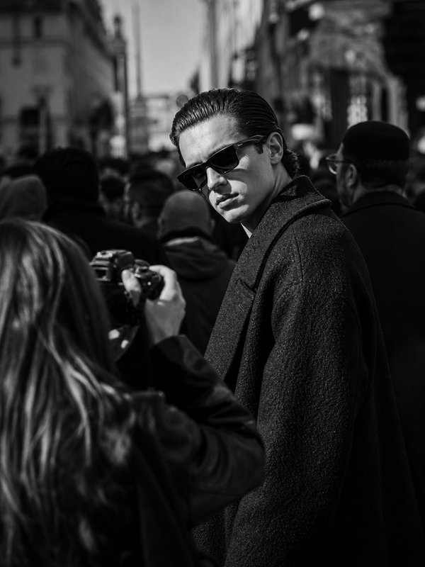 Black and white street photo of stylish man in sunglasses being photographed at a crowded urban fashion event.