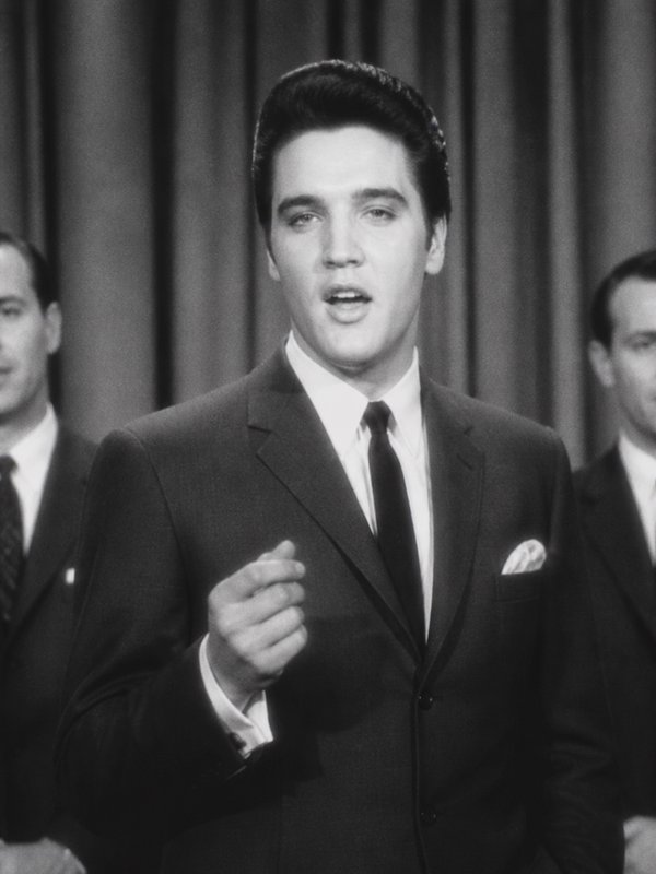 Vintage black and white shot of Elvis Presley performing on a 1960s television stage in a formal tailored suit