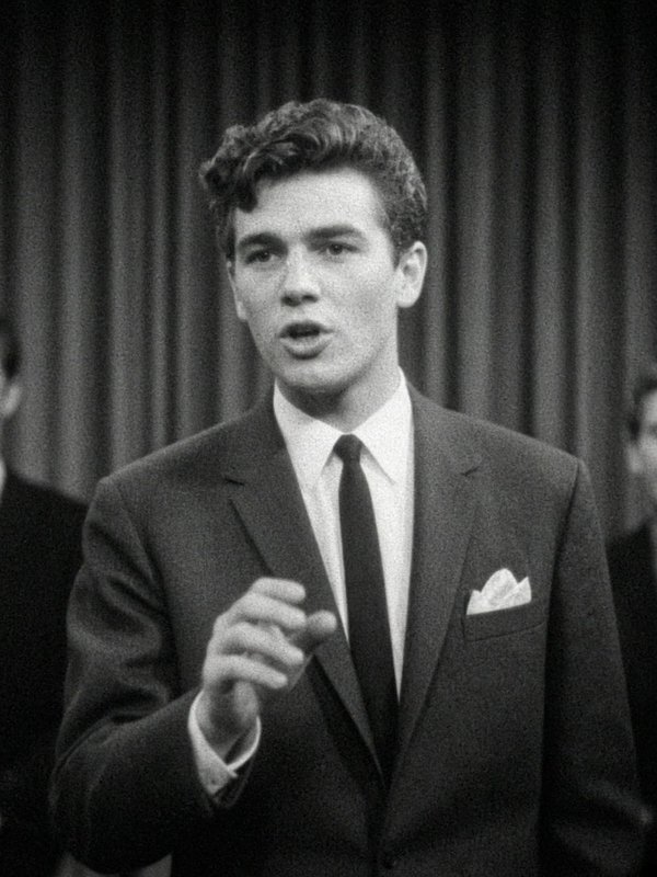 [Black and white vintage shot of young suited man mid-speech, gesturing, stage curtain backdrop behind him]