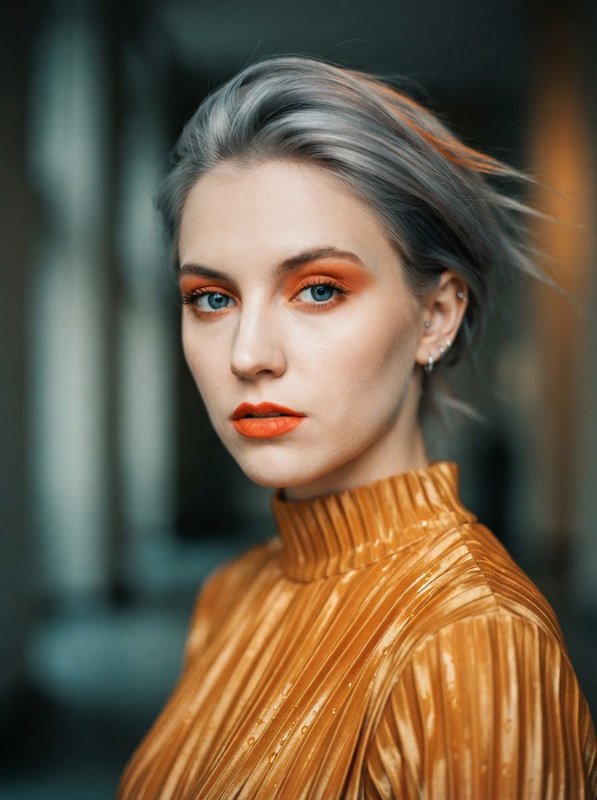 Close-up editorial portrait of a woman with silver hair, bold orange makeup, and a shiny gold pleated turtleneck top