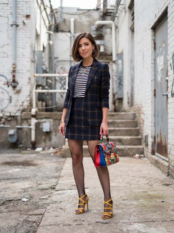 [Fashionable woman in matching plaid blazer skirt set stands in industrial urban alley holding a colorful handbag, wearing...
