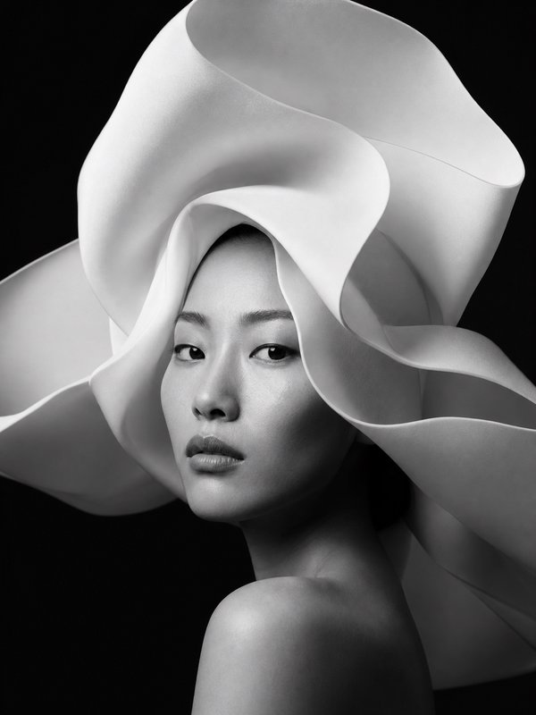 Black and white close-up portrait of elegant East Asian woman wearing large flowing sculptural white headwear against a