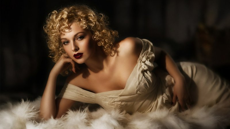 Sultry vintage portrait of a curly blonde woman reclining on fluffy white fur in a cream off-shou...