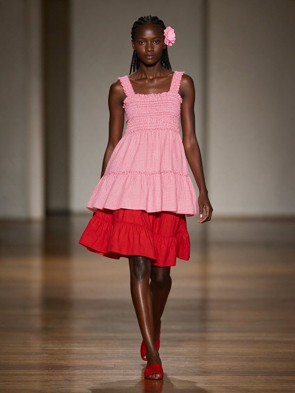 Runway model with braided hair walks in a layered pink gingham top, red ruffled skirt, and pink flower hair accessory.