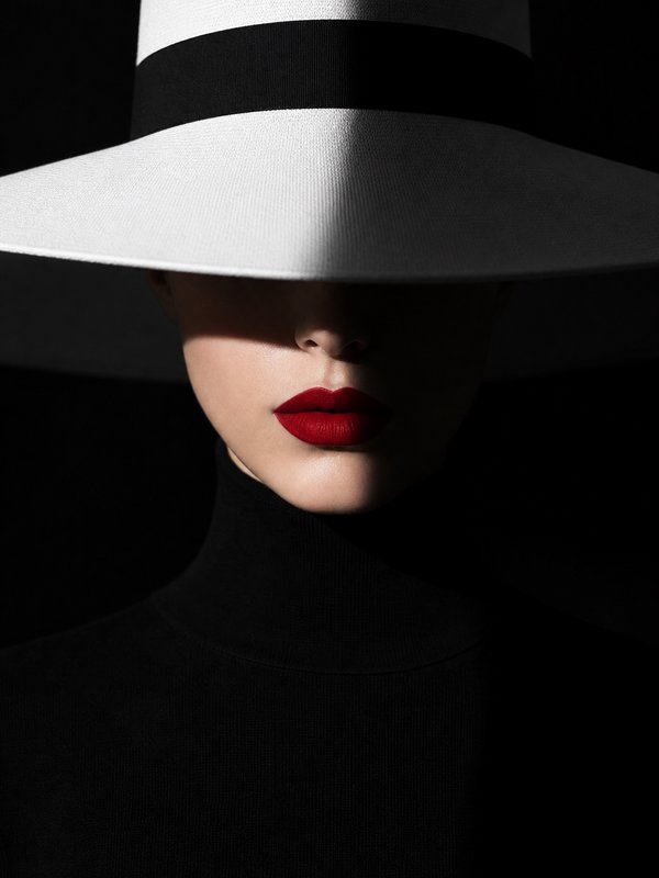 Close-up fashion portrait of a woman with bold red lipstick, her eyes shaded by a white wide-brim hat against a black