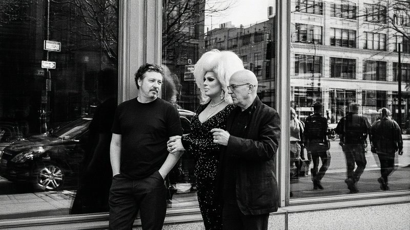 Drag queen with a voluminous bouffant wig stands between two men outside a downtown storefront window