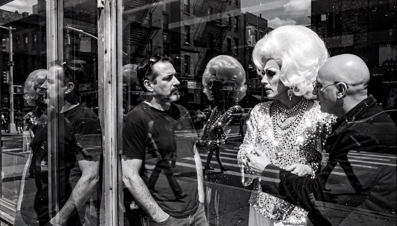 Black and white store window reflection of a glamorous drag queen conversing with two men on a city sidewalk