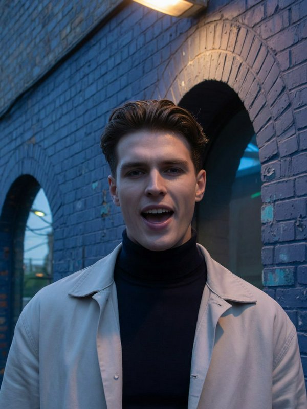 Handsome young man in a turtleneck and overcoat stands in front of a blue brick building at dusk