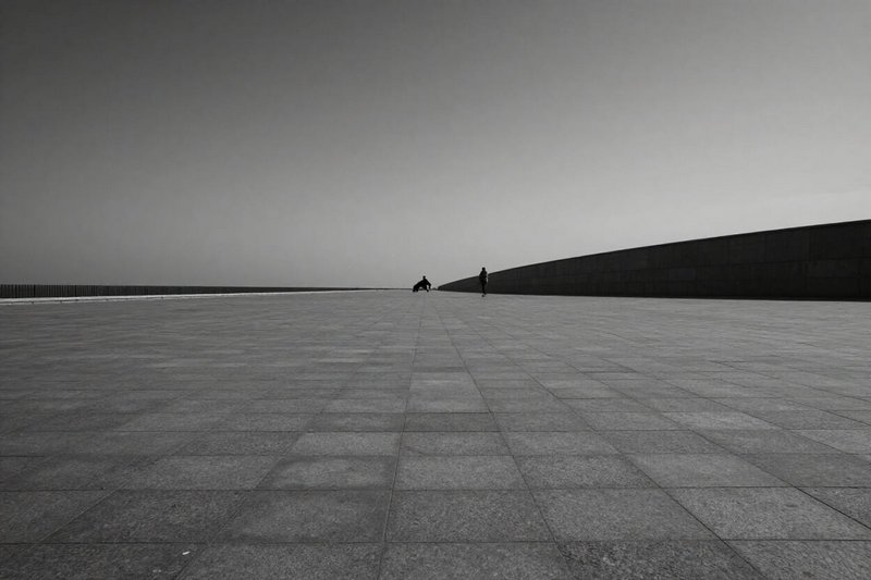 Monochrome image of a vast tiled plaza with a long curved wall and two distant figures.
