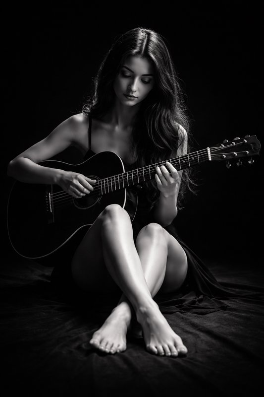 A woman seated on the floor, playing a black acoustic guitar in a moody black-and-white studio po...