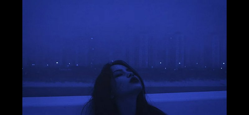 Person with head tilted back in a moody blue night scene against a distant city skyline.