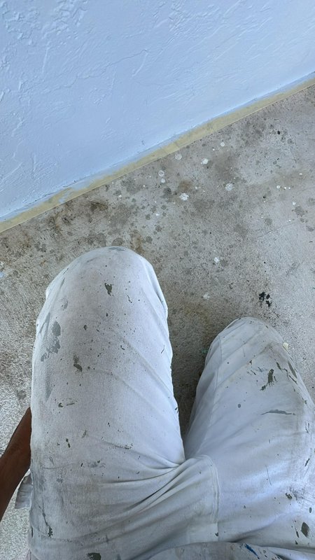 Person wearing paint-splattered white pants kneeling beside a lavender wall on a concrete floor