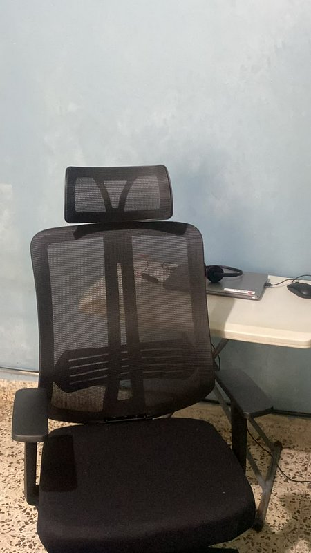 Black mesh ergonomic office chair with headrest next to a small desk holding a laptop, headphones...