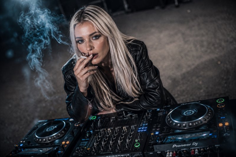 Woman DJ with long blonde hair smoking a cigarette beside a Pioneer DJ mixer.