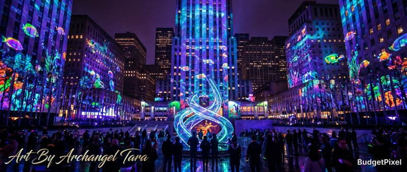 Nighttime city square with colorful building projections and a glowing spiral light sculpture centerpiece.