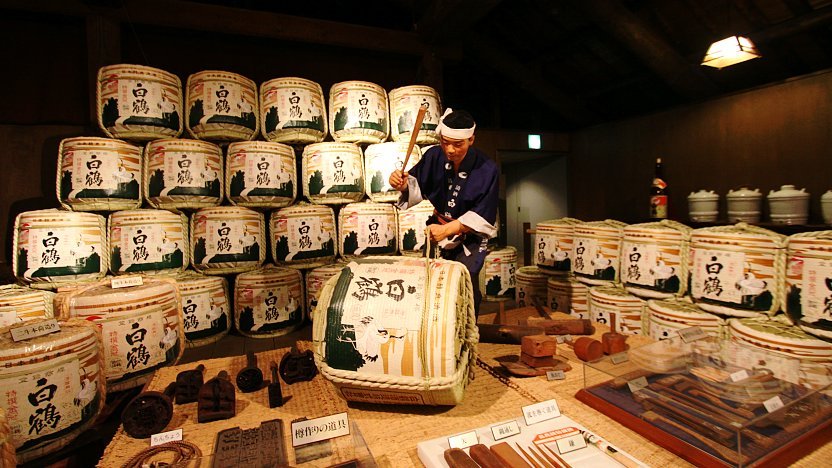 Traditional Japanese sake brewery with stacked wooden barrels and a worker pounding a barrel tapper.