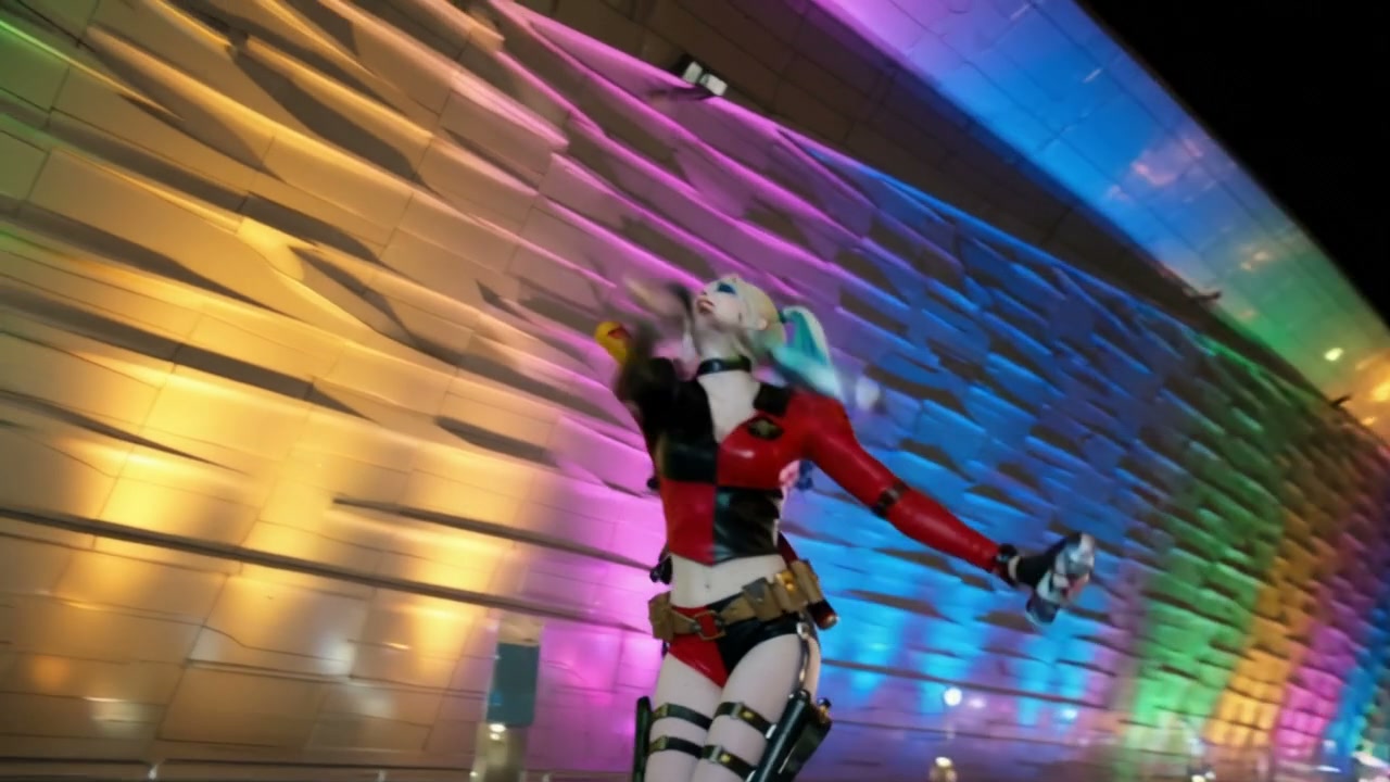 Harley Quinn cosplayer poses playfully at night in front of a modern building lit with vibrant ra...