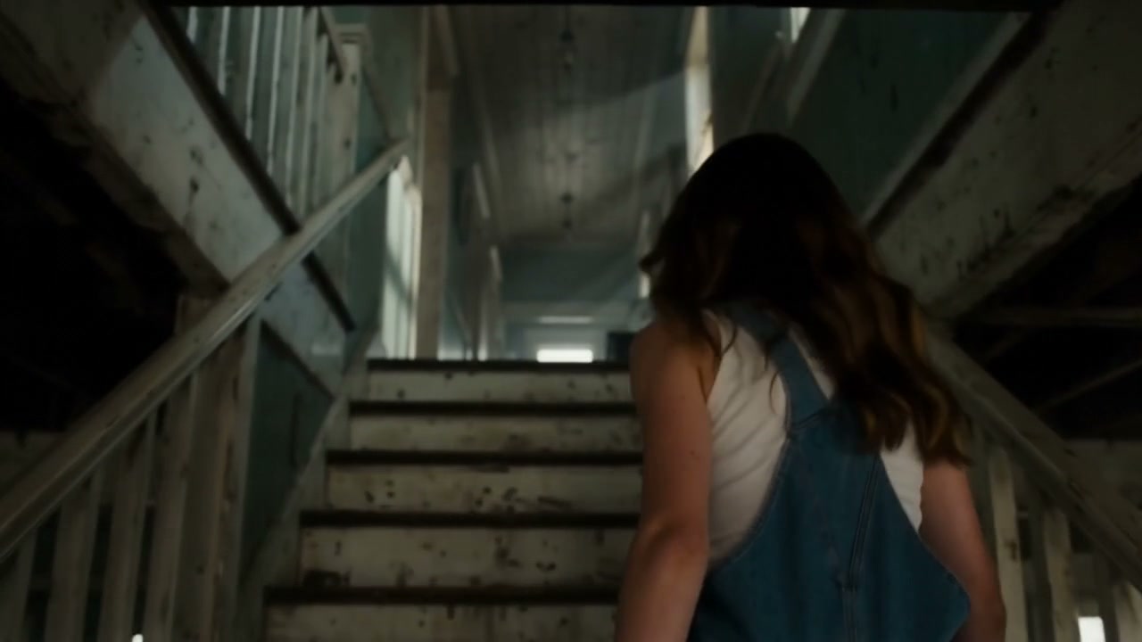 Back view of a young woman climbing the dirty, decaying stairs of an old, abandoned indoor hallway.