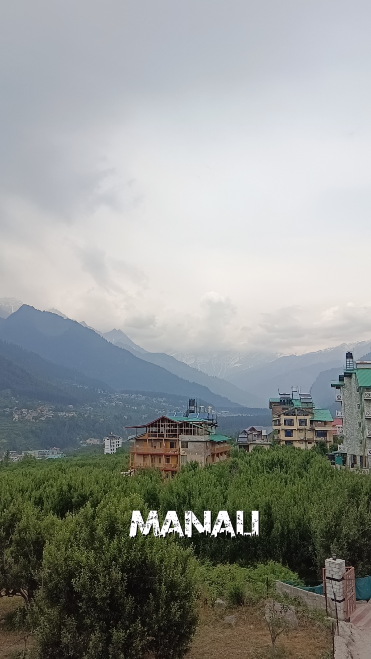 Hillside Manali town with mountains, cloudy sky, and buildings under construction amid green trees.
