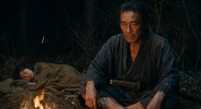 Elderly man in tattered robes sits by a campfire with a katana, while a person sleeps under a bla...