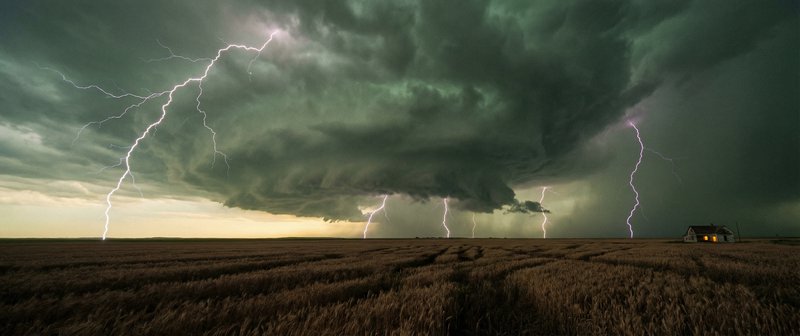 Stormy skies with multiple lightning bolts striking a flat field near a small house.