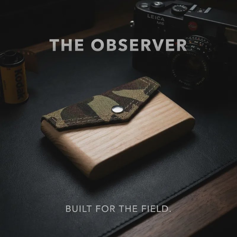 Wooden wallet with camo fabric flap and metal snap, placed on a dark desk with a Leica camera in ...
