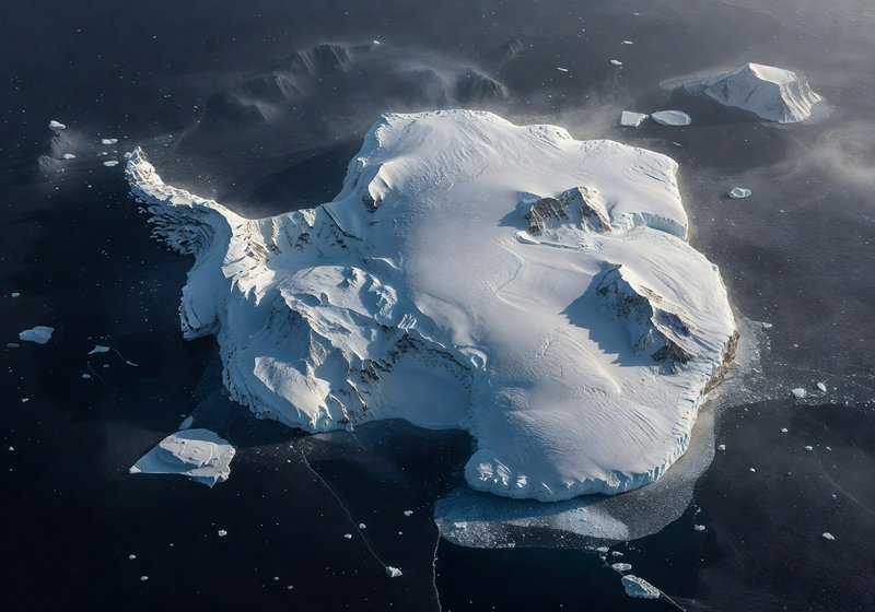 A massive snow-covered iceberg drifts in dark Arctic waters with smaller ice chunks nearby.