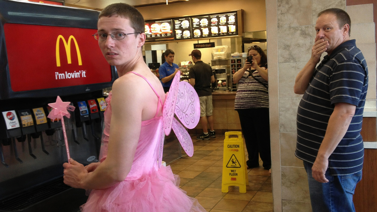 Man in a pink tutu and fairy wings at McDonald's holding a wand near the counter.
