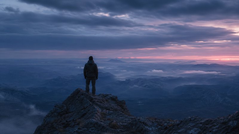 Hiker stands on a jagged mountain peak at sunset, overlooking a misty, blue-toned valley.