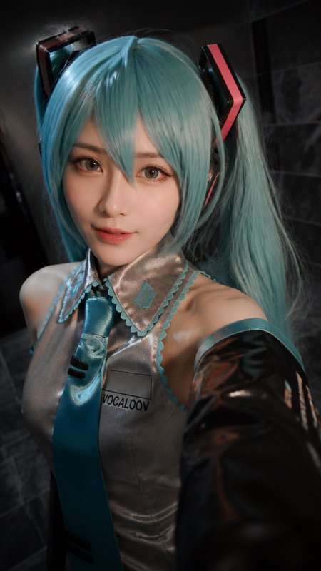 Close-up selfie of a Hatsune Miku cosplayer with turquoise twin tails, wearing the iconic character uniform with a soft ge...