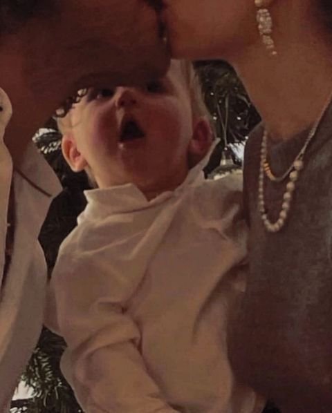 A baby in white clothing with mouth open, held by adults at a family gathering.