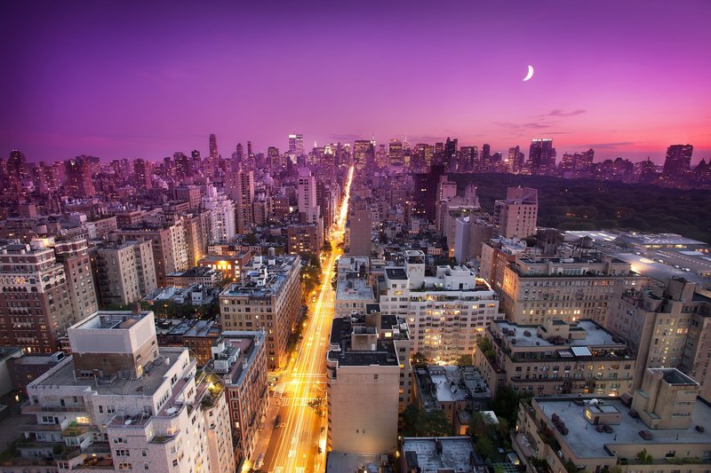 Aerial cityscape at dusk with a glowing main avenue and a crescent moon in a purple sunset sky.