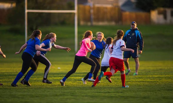 Group of young girls playing rugby on a grassy field during daytime with a coach watching.