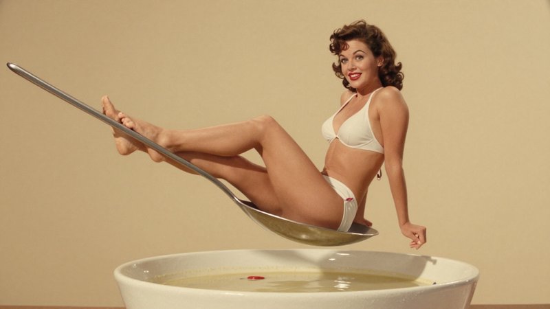 Playful retro pinup woman in a white bikini sits inside an oversized metal ladle hovering above a...