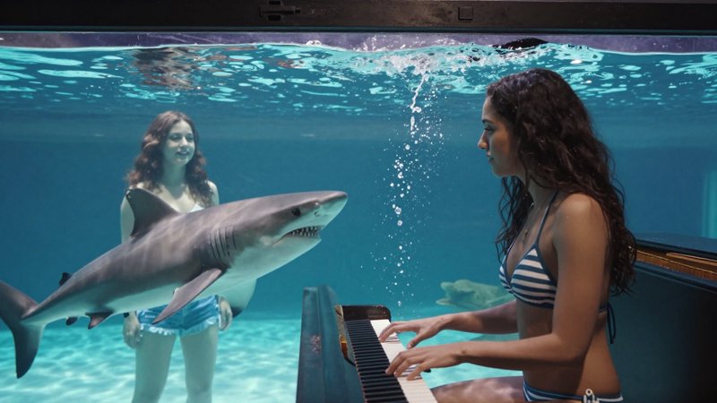 Woman plays piano underwater as a shark and a second swimmer observe in clear turquoise pool water