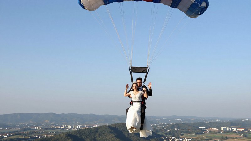 Newlywed bride in a wedding dress and groom wave while descending tandem under a parachute above ...